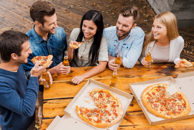 Zorgeloze tijd met vrienden. Bovenaanzicht van vijf vrolijke mensen die flessen bier vasthouden en pizza eten terwijl ze buiten staan