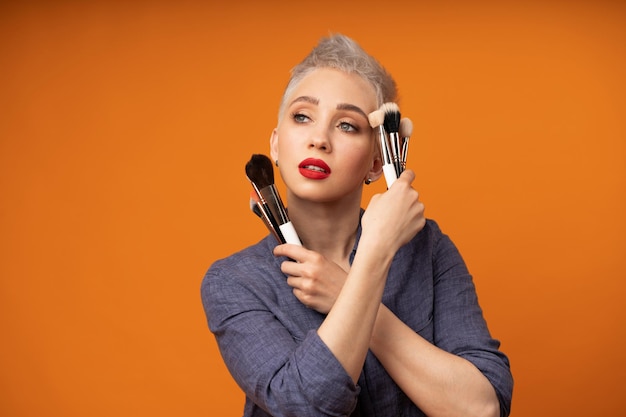 Foto zorgvuldige jonge vrouw met make-upborstels die tegen een oranje achtergrond staat