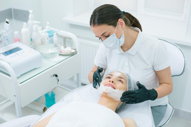 Zorgvuldige schoonheidsspecialist in rubberen handschoenen en medisch masker die zuurstofmasker toepast op een gezicht van haar cliënt