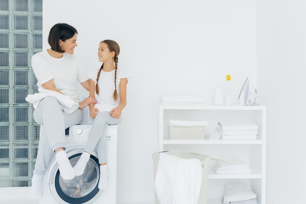 Zorgzame moeder praat met dochter pose op wasmachine omringd met wit linnen wassen samen in de wasruimte thuis gelukkig volwassen huisvrouw bedankt schattig meisje voor hulp