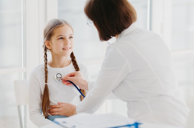 Zorgzame vrouwelijke kinderarts luistert naar de longen of het hart van het kind met een fonendoscoop.