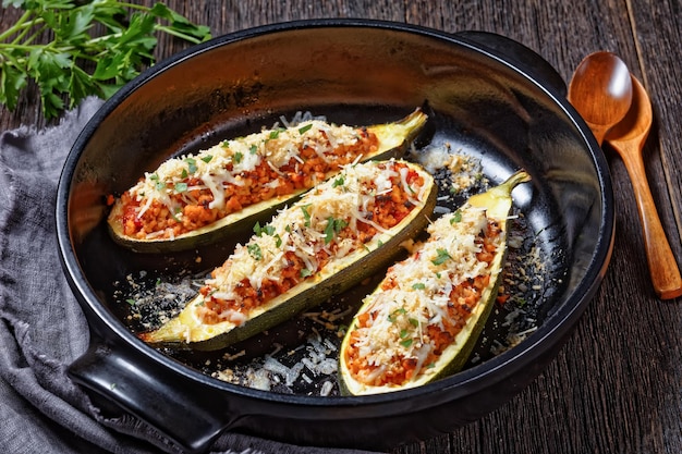 Barche di zucchine ripiene di carne macinata, pomodori tritati e formaggio in una teglia nera su un tavolo di legno, cucina italiana