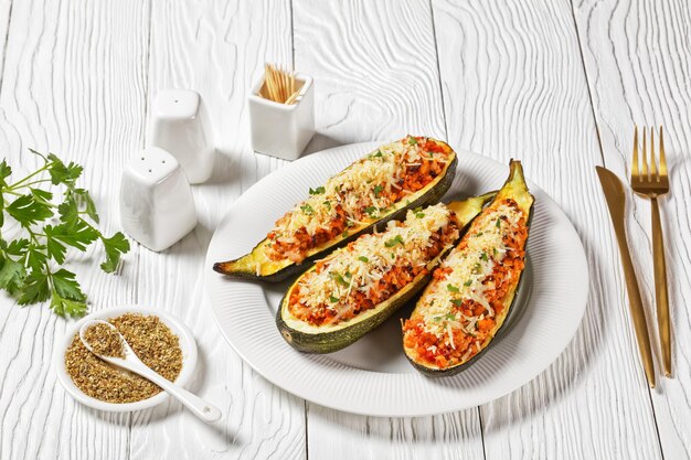 Zucchini boats stuffed with ground meat, chopped tomatoes, and cheese on a white platter on a wooden table,  italian cuisine