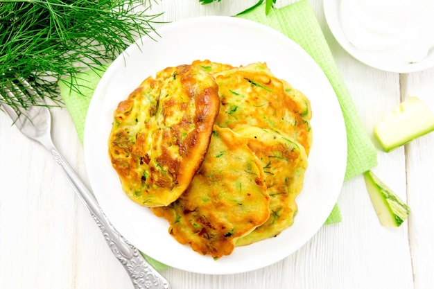 Zucchini fritters, dill and parsley in a plate on a napkin, sour cream in platter on wooden board background from above