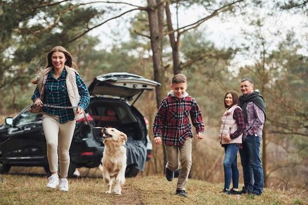 Zus en broer rennen vooruit Gelukkig gezin veel plezier met hun hond in de buurt van moderne auto buiten in het bos