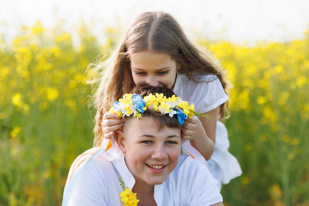 Zus vlecht linten in Oekraïense krans met bloemen op hoofd van oudere broer op weide tegen veld