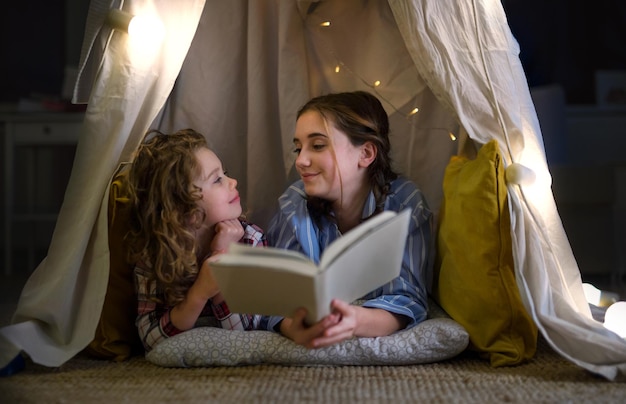 Zusters binnenshuis thuis, leesboek in pyjama in tent. Lockdown-concept.