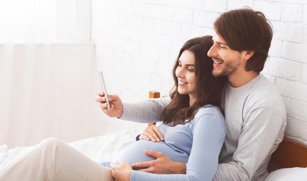 Zwanger echtpaar ontspant zich thuis met een smartphone op het bed
