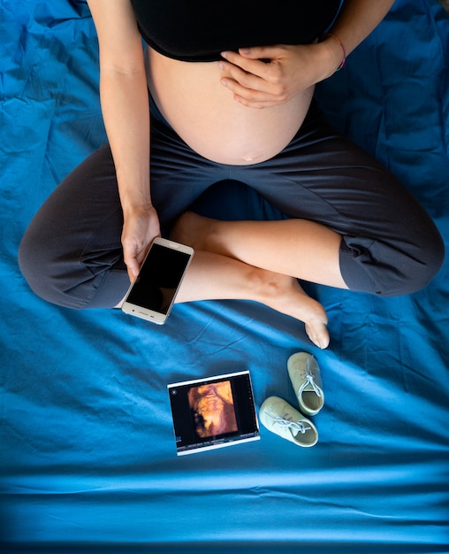 Foto zwanger meisje dat de telefoon met babyschoenen en ecography-zitting op een blauw bed bekijkt
