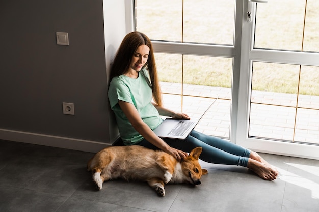 Zwanger meisje met laptop glimlach en speel thuis met corgi-hond programmeur vrouw werkt online en heeft een pauze om haar hond te knuffelen plezier hebben met welsh corgi pembroke