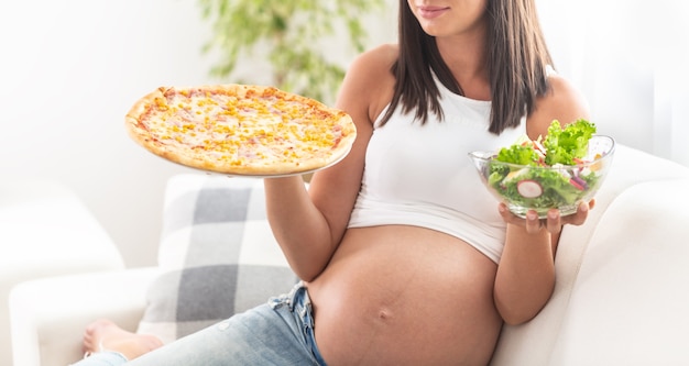 Zwanger meisje zittend op een bank met slakom in de ene hand en pizza in de andere.