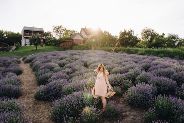 Zwanger meisjesblonde in een beige kleding en een strohoed. lavendel veld. in afwachting van een kind. het idee van een fotoshoot. loop bij zonsondergang. toekomstige moeder. mand met bloemen.