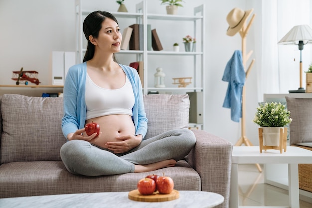 Zwangere aziatische vrouw houdt van een gezonde maaltijd met fruit- en groentesalade om voor de baby in de baarmoeder te zorgen om sterk te zijn. jonge mooie lachende moeder met verse rode appel zittend in de bank kijkvenster