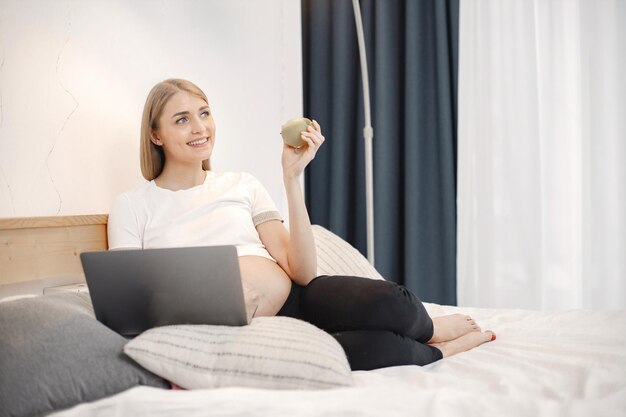 Zwangere dame ligt op een bed in de kamer en houdt een appel vast terwijl ze een laptop gebruikt