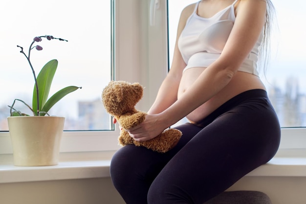 Zwangere jonge gelukkige vrouw met speelgoedteddybeer thuis in de buurt van raam