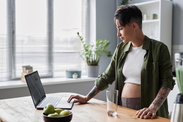 Zwangere jonge vrouw die laptop gebruikt die zich in comfortabele keuken bevindt