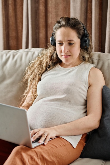 Zwangere jonge vrouw in koptelefoon die op zijn gemak naar online video kijkt