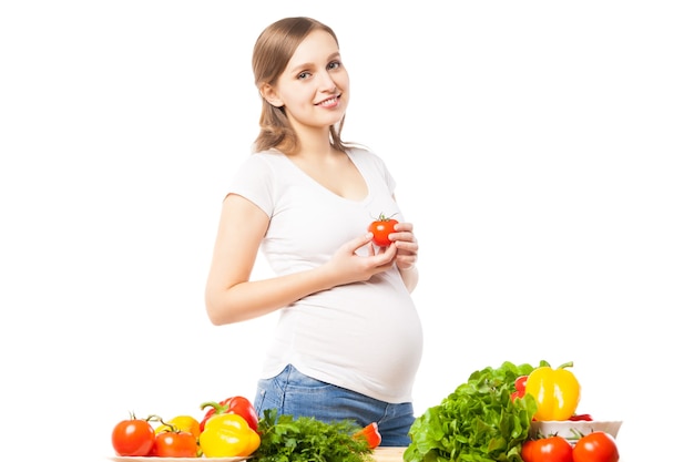 Zwangere lachende vrouw met tomaat aan tafel vol groenten. Studio shot