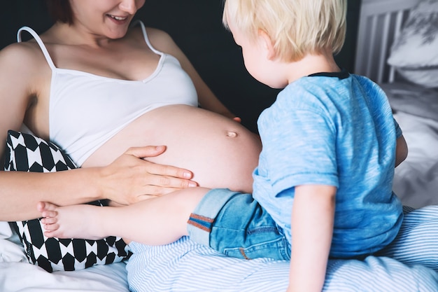 Zwangere moeder en zoon samen in bed thuis Klein kind dat naar de buik van haar moeder kijkt