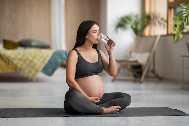 Zwangere mooie vrouw drinkt water uit glas terwijl ze thuis yoga beoefent