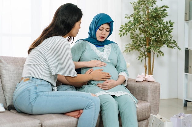 Zwangere moslimvrouw die buikpijn heeft en een vriend die haar troost op de comfortbank in de woonkamer. ziekte kraamvrouw met hoofddoek pijnlijk gevoel op buik. toekomstige moeder met pijnlijke buik