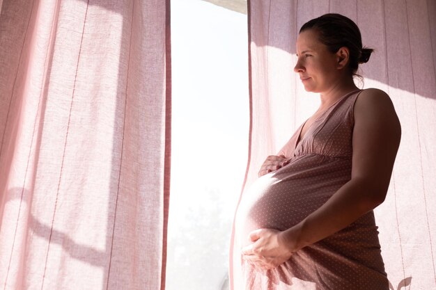 Zwangere vrouw die de buik aanraakt die bij het raam staat