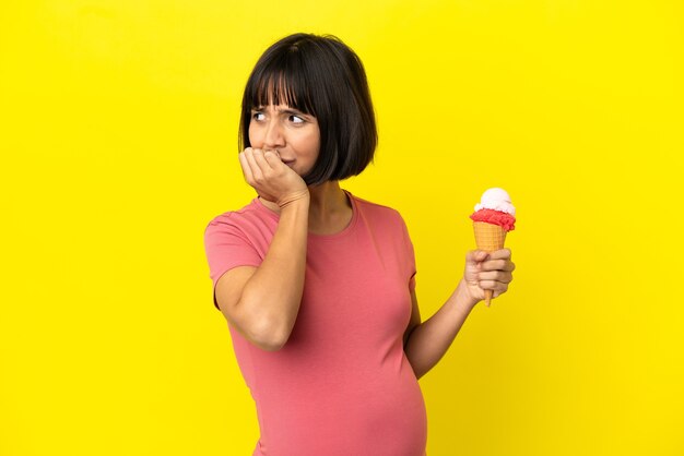 Zwangere vrouw die een cornetroomijs houdt dat op gele achtergrond wordt geïsoleerd en twijfels heeft