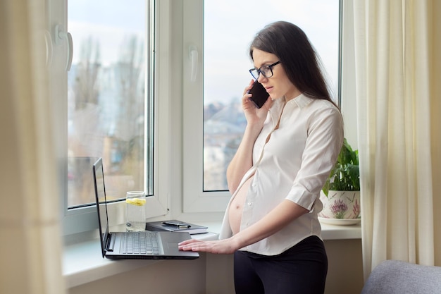 Zwangere vrouw die op afstand thuis werkt. Jonge zakenvrouw met laptop praten aan de telefoon bij het raam.