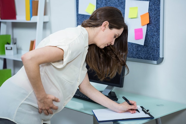 Zwangere vrouw die op klembord bij bureau schrijft