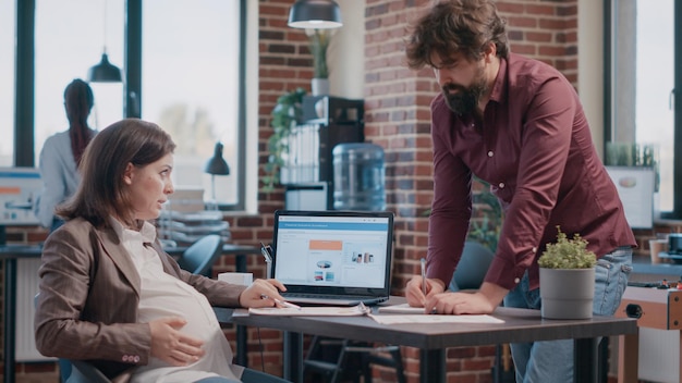 Zwangere vrouw die samenwerkt met collega voor zakelijk project. Team van zakenmensen die samenwerken aan startup-ontwikkeling, met behulp van papieren en laptop om marketingstrategie te ontwerpen.