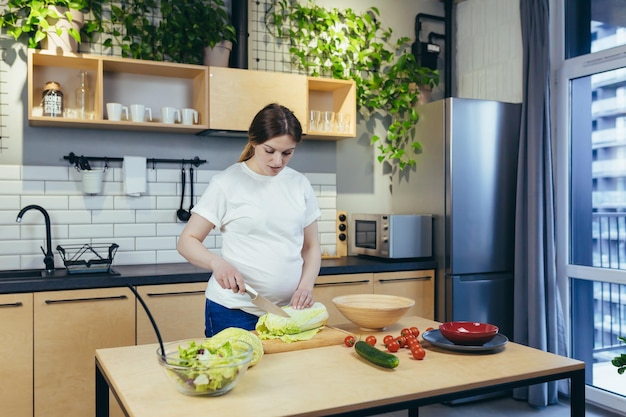 Zwangere vrouw die thuis in de keuken gezond voedsel voorbereidt, snijdende groenten voor salade