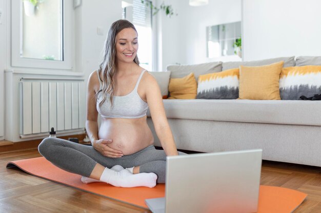 Zwangere vrouw die thuis yoga beoefent met laptop. Aanstaande moeder doet prenatale videotraining binnenshuis. Vrouwelijke oefening, mediteer tijdens de zwangerschap. Online fitnessles op digitale apparaten.