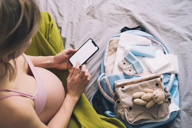 Foto zwangere vrouw die zich voorbereidt op de bevalling, spullen inpakt voor het ziekenhuis, aantekeningen maakt of een checklist in de smartphone, babykleding, benodigdheden voor moeder en pasgeborene in de kraamzak