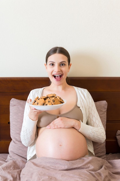 Zwangere vrouw geniet van het eten van koekjes die thuis in bed rusten Ongezond zoet gebak tijdens de zwangerschap concept