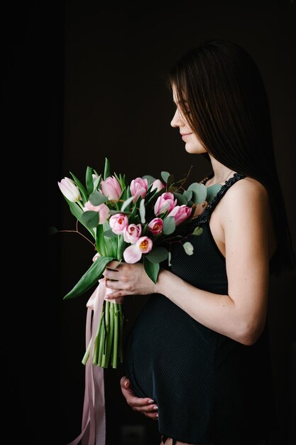 Zwangere vrouw houdt boeket bloemen in handen meisje omarmt een ronde buik in de buurt van het raam thuis permanent zijwaarts moederschap concept close-up negen maanden baby shower