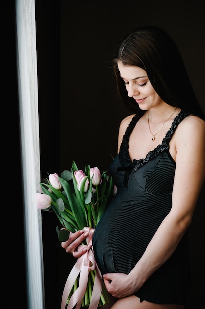 Zwangere vrouw houdt boeket bloemen in handen meisje omarmt een ronde buik in de buurt van het raam thuis permanent zijwaarts moederschap concept close-up negen maanden baby shower