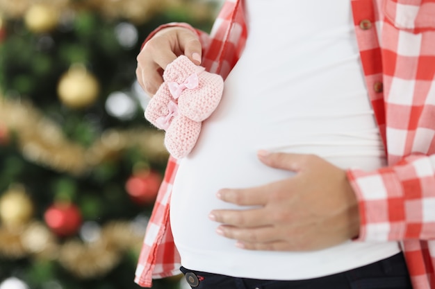 Zwangere vrouw houdt roze babyslippers vast op de achtergrond van nieuwjaarsboom kerstmis en moederschap