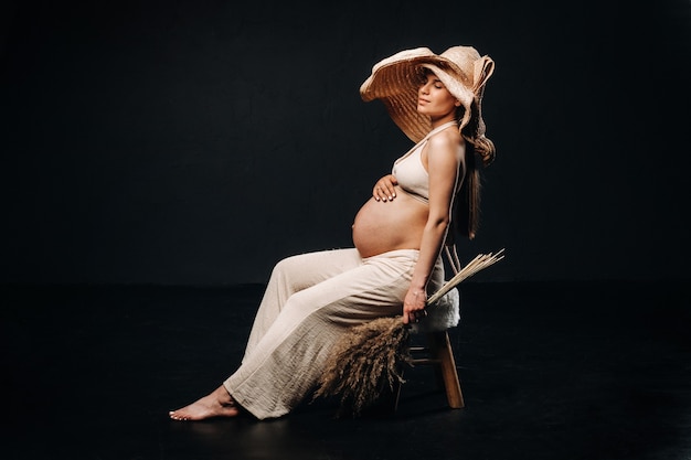 Zwangere vrouw in een strohoed met beige kleding met een boeket in haar handen in de studio op een zwarte achtergrond.