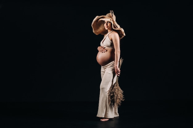 Zwangere vrouw in een strohoed met beige kleding met een boeket in haar handen in de studio op een zwarte achtergrond.