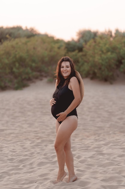 zwangere vrouw in een zwart zwempak en hoed op een zandstrand