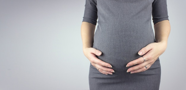 Zwangere vrouw in jurk houdt handen op buik op een grijze achtergrond. Moeder verwachting.