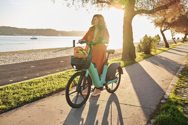 Zwangere vrouw in oranje jas op elektrische fiets op de promenade bij zonsondergang