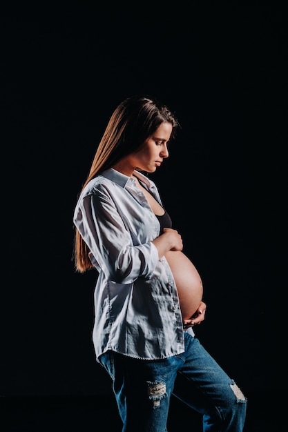 Zwangere vrouw in wit overhemd en jeans in studio op zwarte achtergrond.