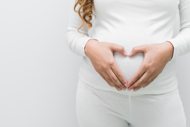 Zwangere vrouw in witte jurk maakt handvorm hart op haar buik op witte backgroundasian woman