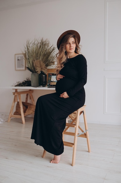 Zwangere vrouw in zwarte jurk zittend op de stoel in de studio met witte achtergrond Zwangere vrouw die buik aanraakt