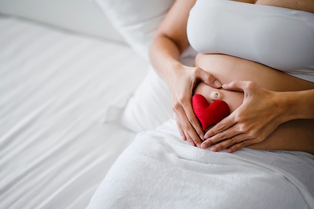 Zwangere vrouw is blij om voor het kind te zorgen bij de geboorte. Gelukkige zwangere vrouw raakt haar buik aan. Zwangere vrouw toont liefde door een hartvorm op hun buik te maken.