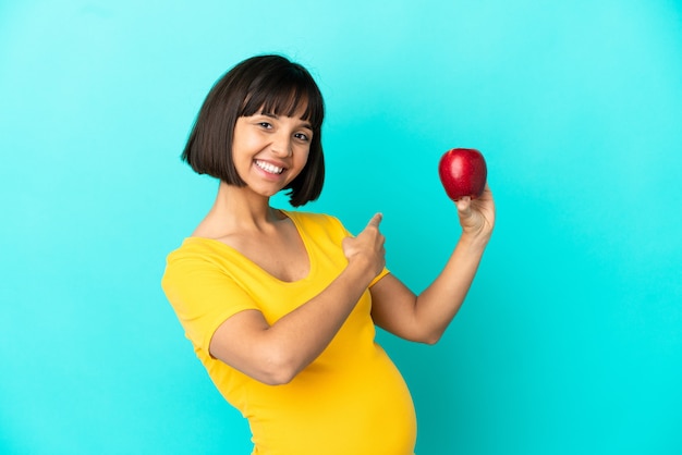 Zwangere vrouw met een appel geïsoleerd op een blauwe achtergrond die terug wijst