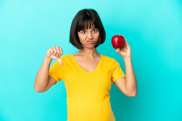 Zwangere vrouw met een appel geïsoleerd op een blauwe achtergrond met duim omlaag met negatieve uitdrukking