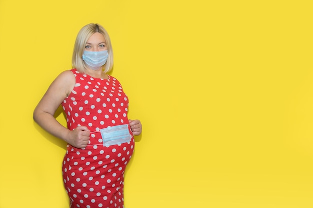 Zwangere vrouw met een masker houdt een masker in haar handen en bedekt haar buik op een gele achtergrond met kopieerruimte