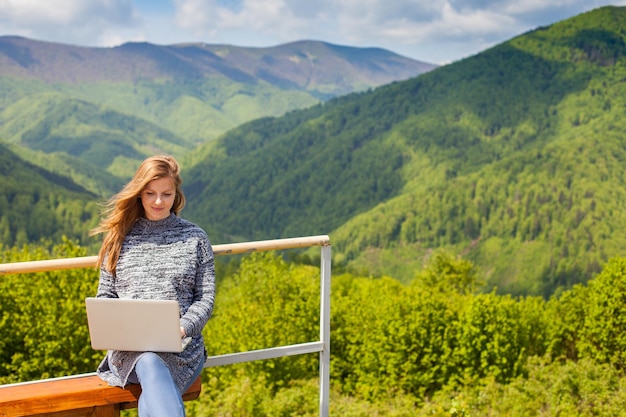 Zwangere vrouw met lang haar zoekt iets in haar laptop op een houten bankje op de achtergrond van de natuur